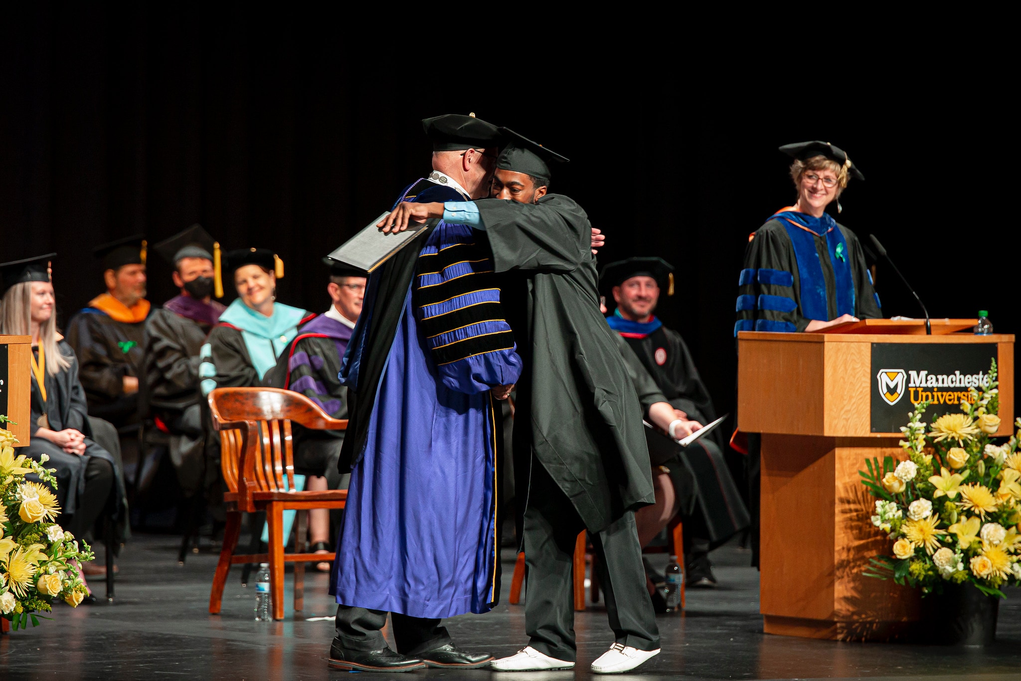 Commencement student walking on stage hugging faculty Commencement student walking on stage hugging faculty