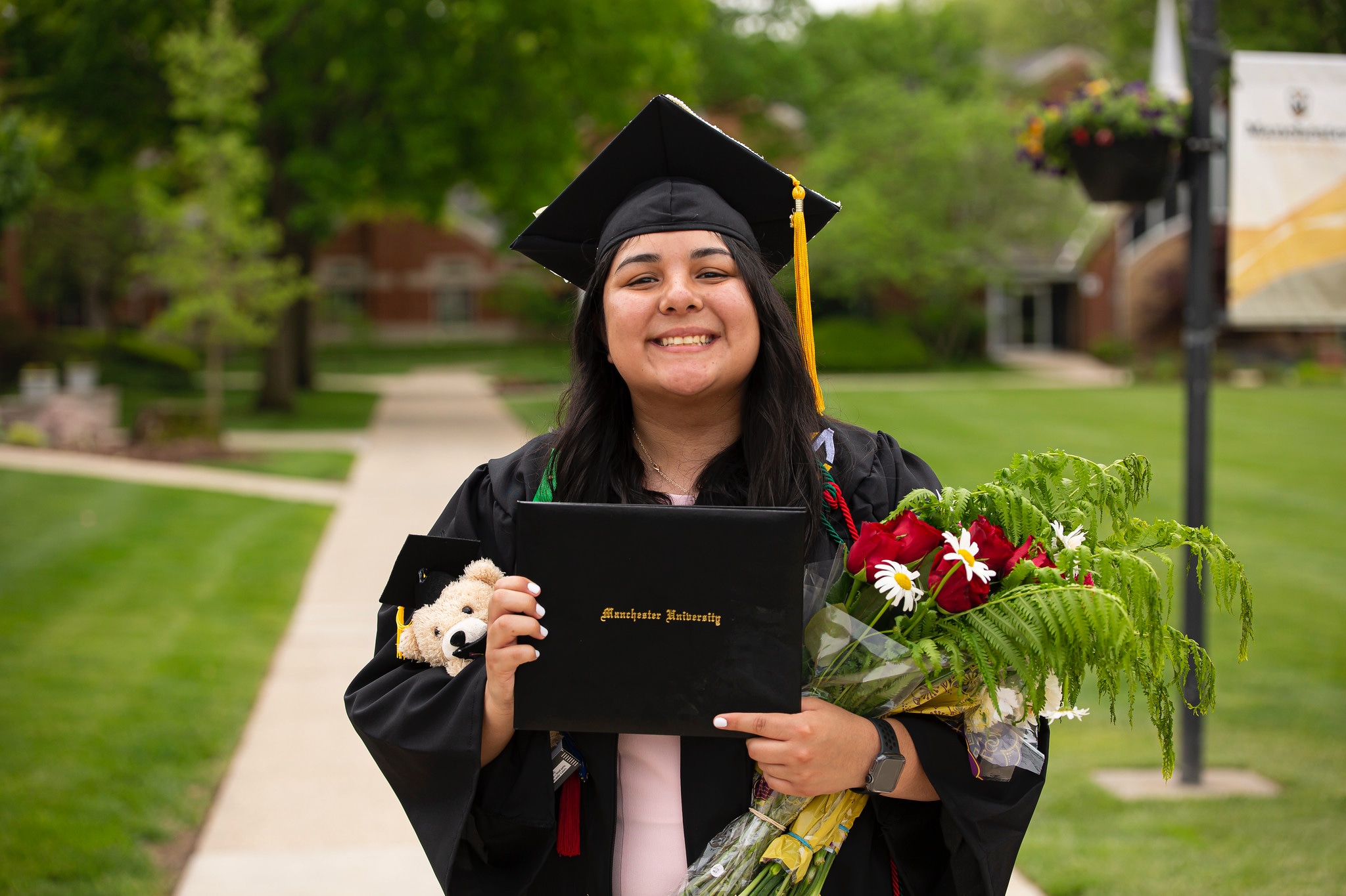 Commencement student with flowers and teddy bear outside smiling Commencement student with flowers and teddy bear outside smiling