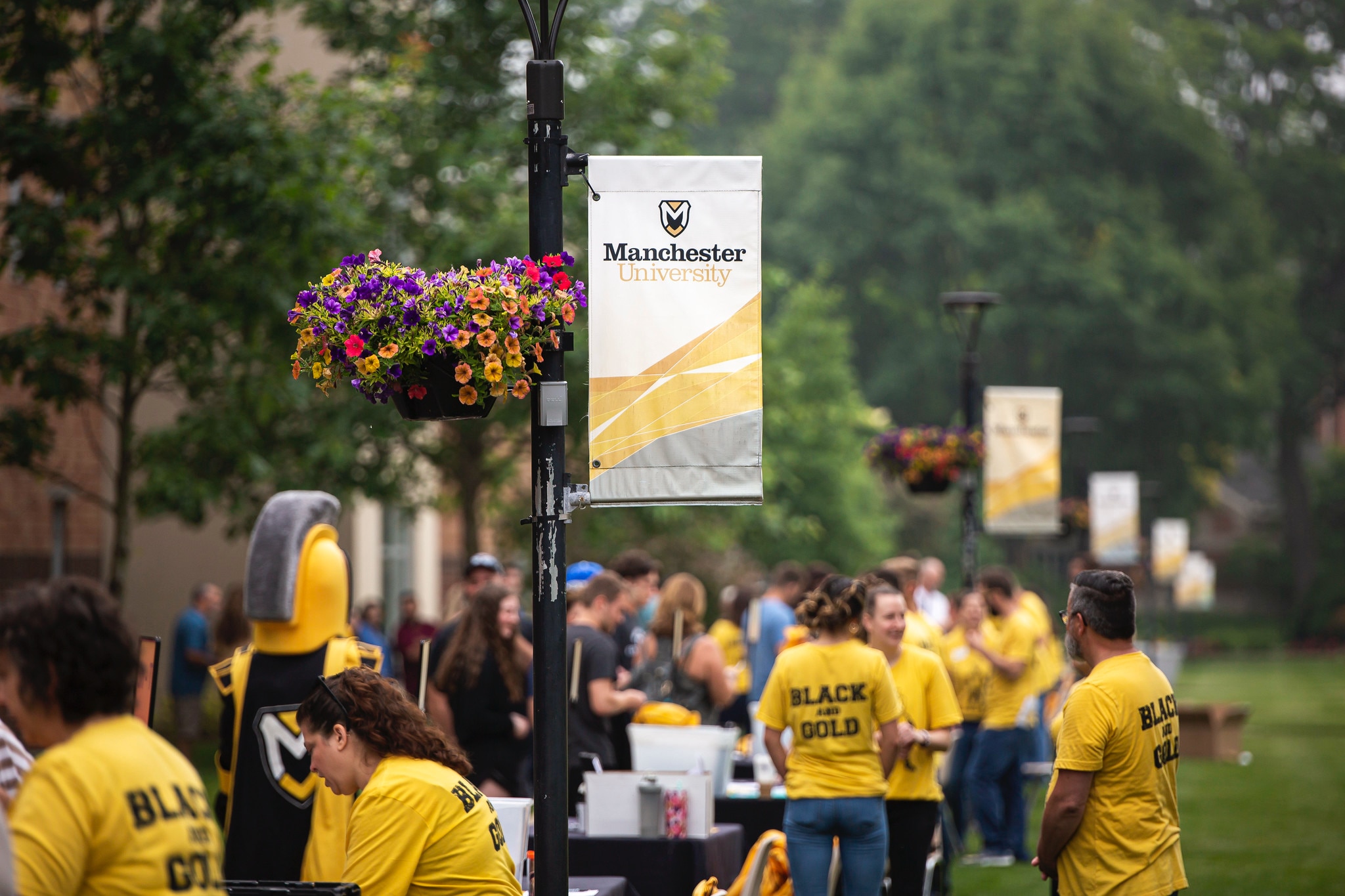 Black and Gold Visit Day students at table with pole sign of logo Black and Gold Visit Day students at table with pole sign of logo