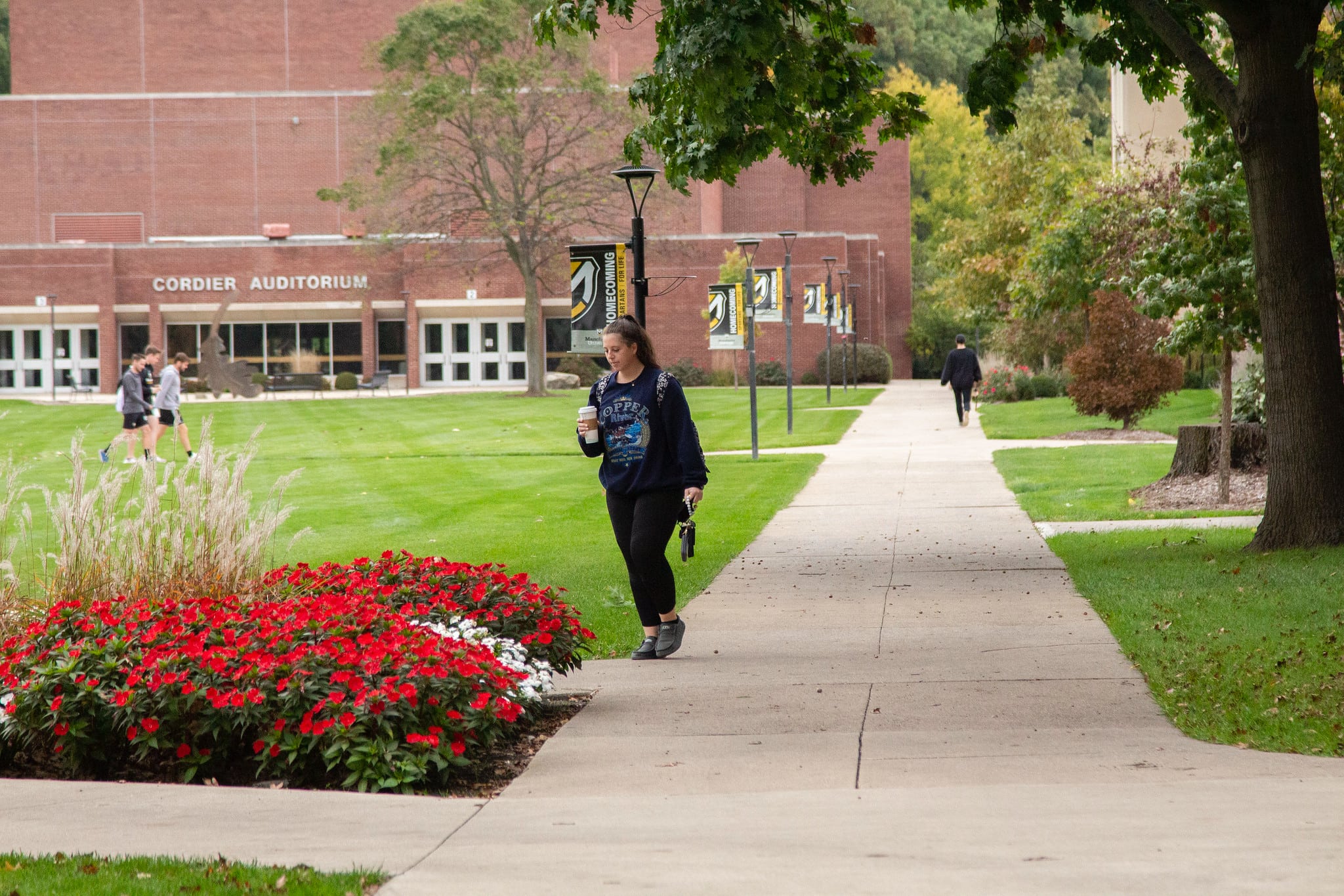 student walking on campus with coffee in fall with red flowers wiht Cordier Auditorium in background