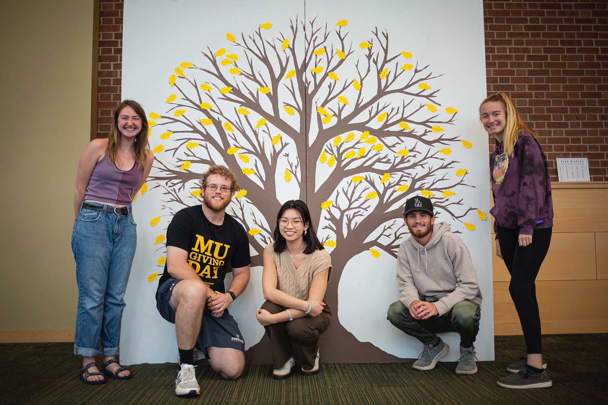 students inside a building posing for a picture in front of a painted tree