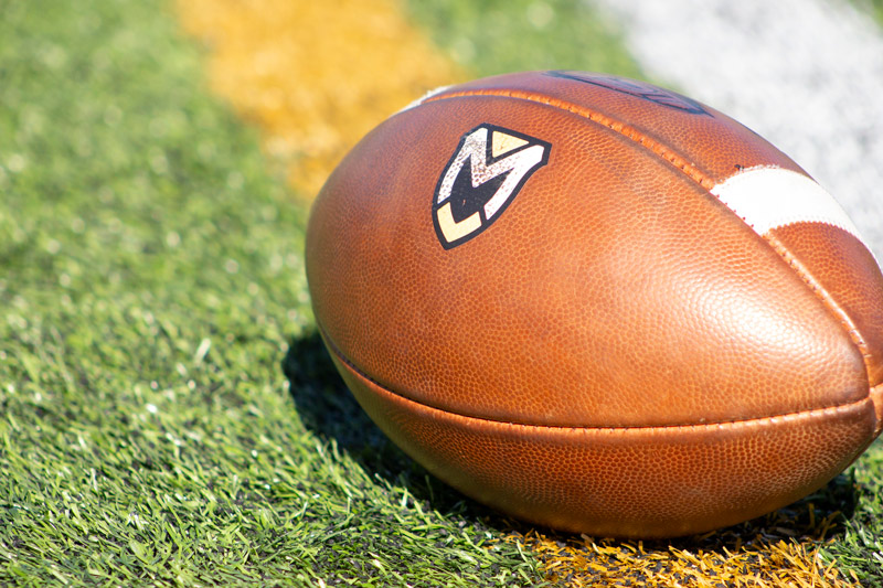 Image is a closeup of an MU branded football that displays the MU Logo on the football field turf