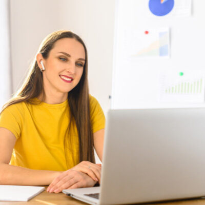 Smiling woman with wireless earbuds sits at desk with laptop, notebook and pen in white office.