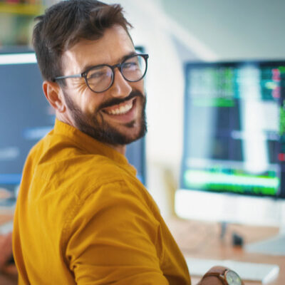 Smiling man with beard and glasses looking back over right shoulder while sitting infrom of a computer screen, while a person sitting to his left works at their computer.
