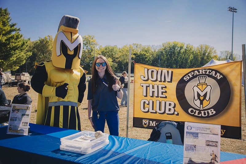 A sign reads Join the Club for the Spartan Club with Manny the Spartan standing beside a sign up booth.