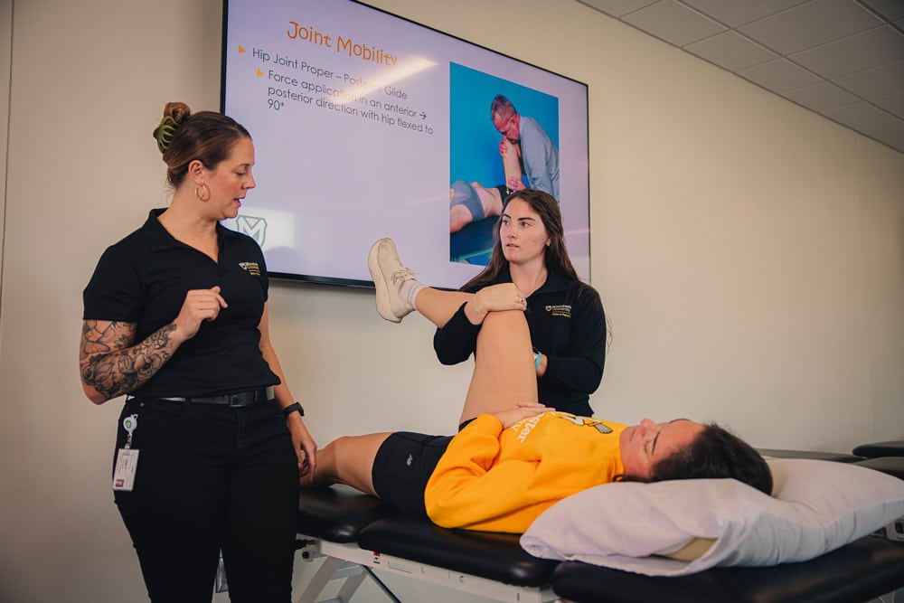 A student works under the instruction of a professional in the MU Fort Wayne Physical Therapy lab while stretching the leg of a female volunteer in a yellow MU hoodie.