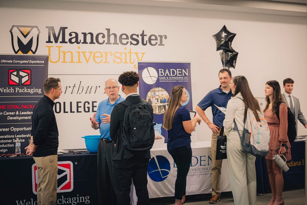 Multiple students ineract with local area small and large business leaders at a College of Business mixer. Vendor tables are nearby with MU logo prominently placed in the background.