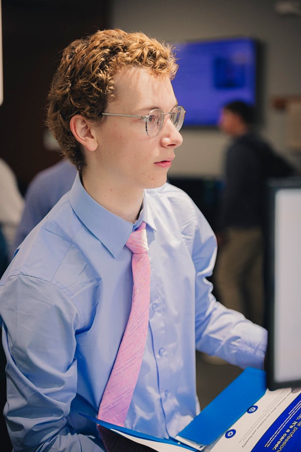 A male student with curly hair and a pink tie against a blue dress shirt attends an MBA mixer. 