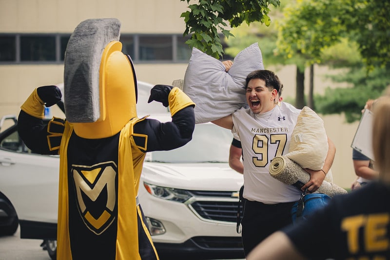 Manny the mascot flexes his muscles while a student carries a pillow making a joke.