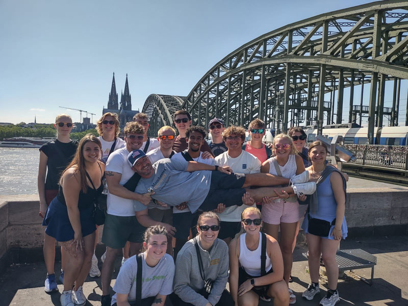 Students hold Dr. Jeff Beer horizontally with the bridge from Cologne Germany in the background. 