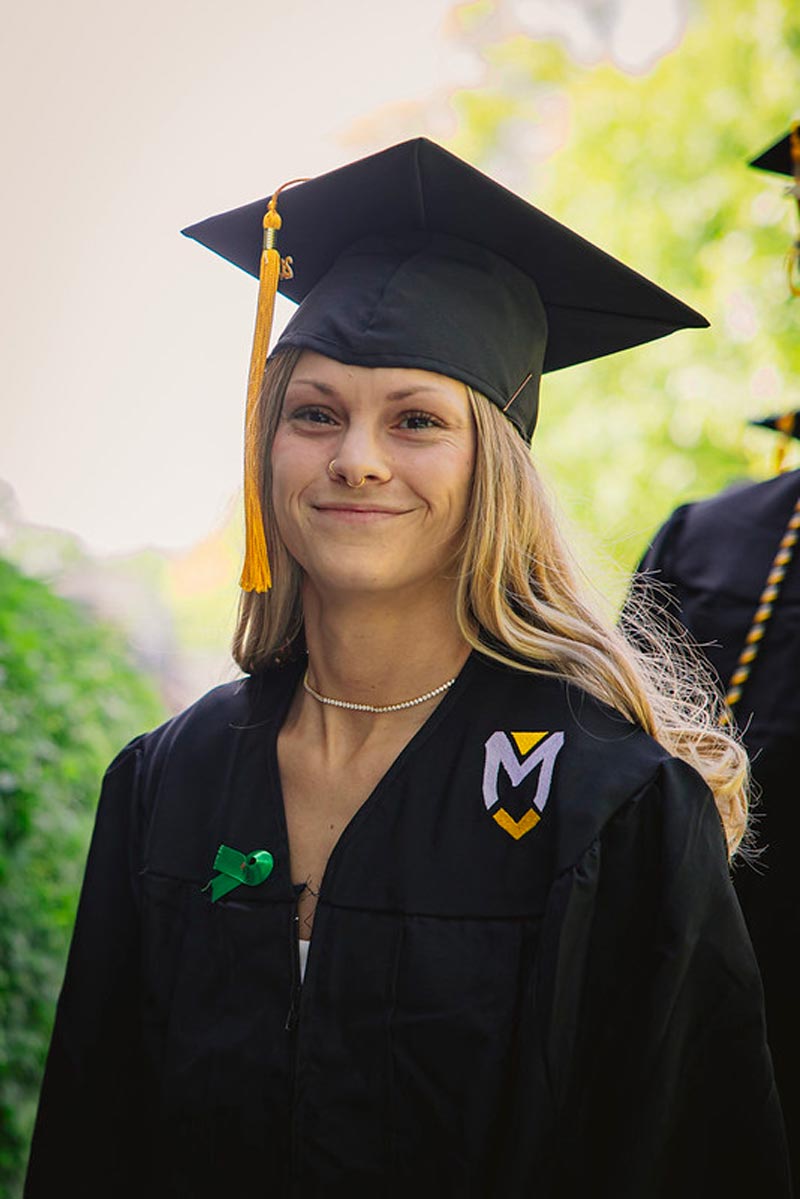 A female student in graduation cap and gown, wearing a green ribbon, smiles at the camera