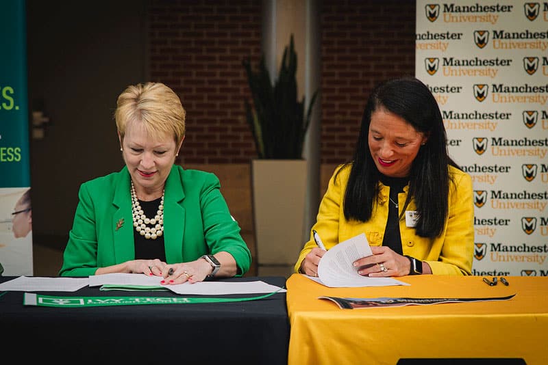 Ivy Tech and Manchester University leadership sign an agreement at respective tables decorated in their school colors. 