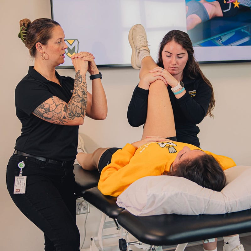 An instructor oversees the process within the PT Lab by giving direction to a student and a volunteer providing stretching exercises