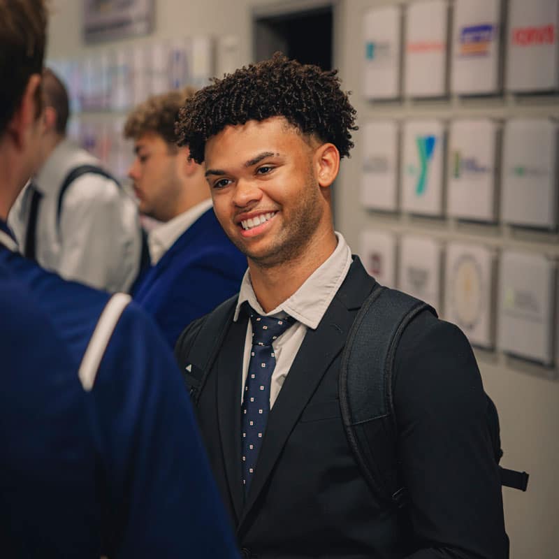 Smiling in conversation, a male student with a backpack wears a suit.
