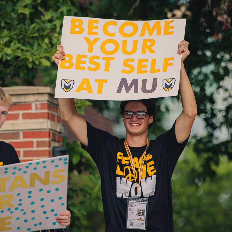 A student lifts a celebratory poster over his head that reads "Become your best self at MU"