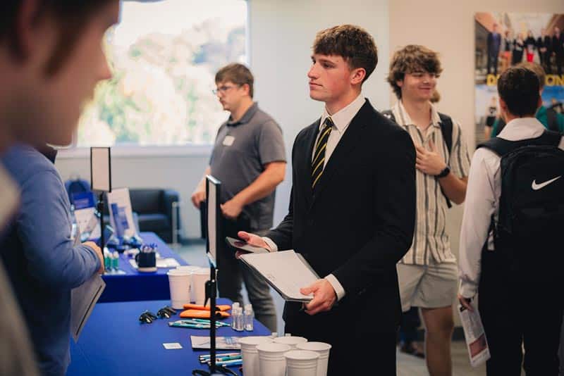 A Manchester student is at a small table at a convention. 