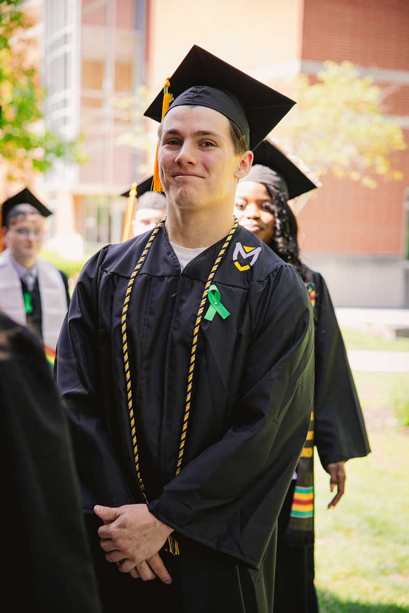 A male student walks to receive his unergrad degree wearing a black cap and gown and green ribbon. 