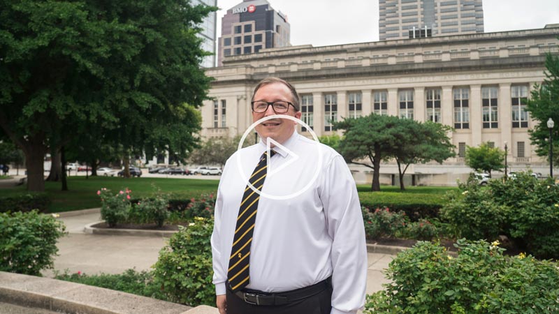Josih Hostetler stands in front of a larger building where he works, a play video thumbnail rests over the image - click the image to play the video.