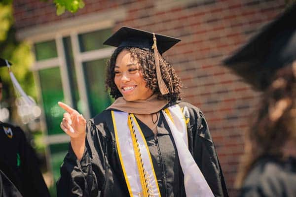 A female graduate points and smiles dressed in cap and gown.