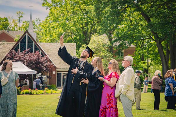 A young man in cap and gown takes a selfie with his family at graduation
