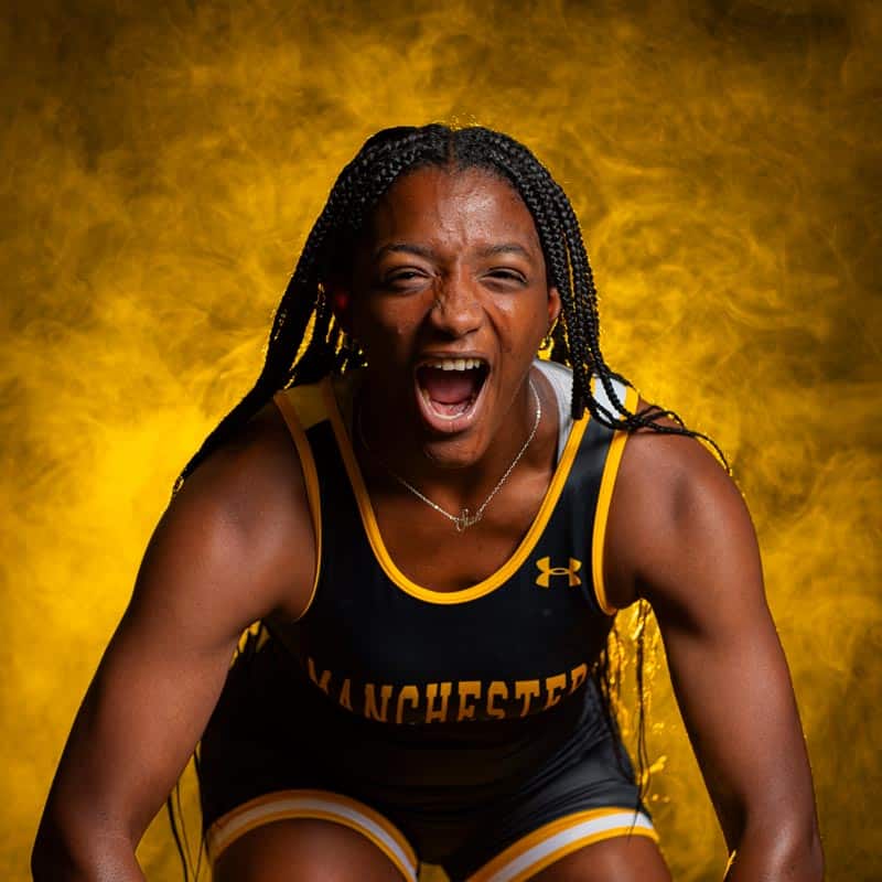 Female-Wrestler-Backlit-Fog Manchester University Division III Women's Wrestling female athlete flexing her muscles and fiercely showing her strength.