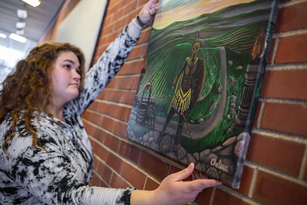A Manchester University student hangs a gallery painting of a spartan standing in a field in full armor.