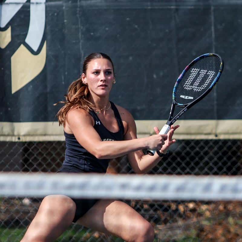 A Manchester female tennis player chases after a backhand return with the Spartan logo behind her.