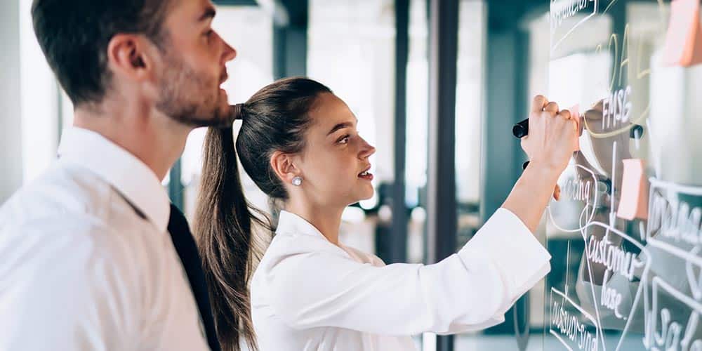 2 Team members stand side by side in front of a whiteboard made of clear glass and create plans.
