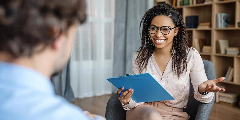 An instructor sitting with a clipboard smiling at an onlooking student.