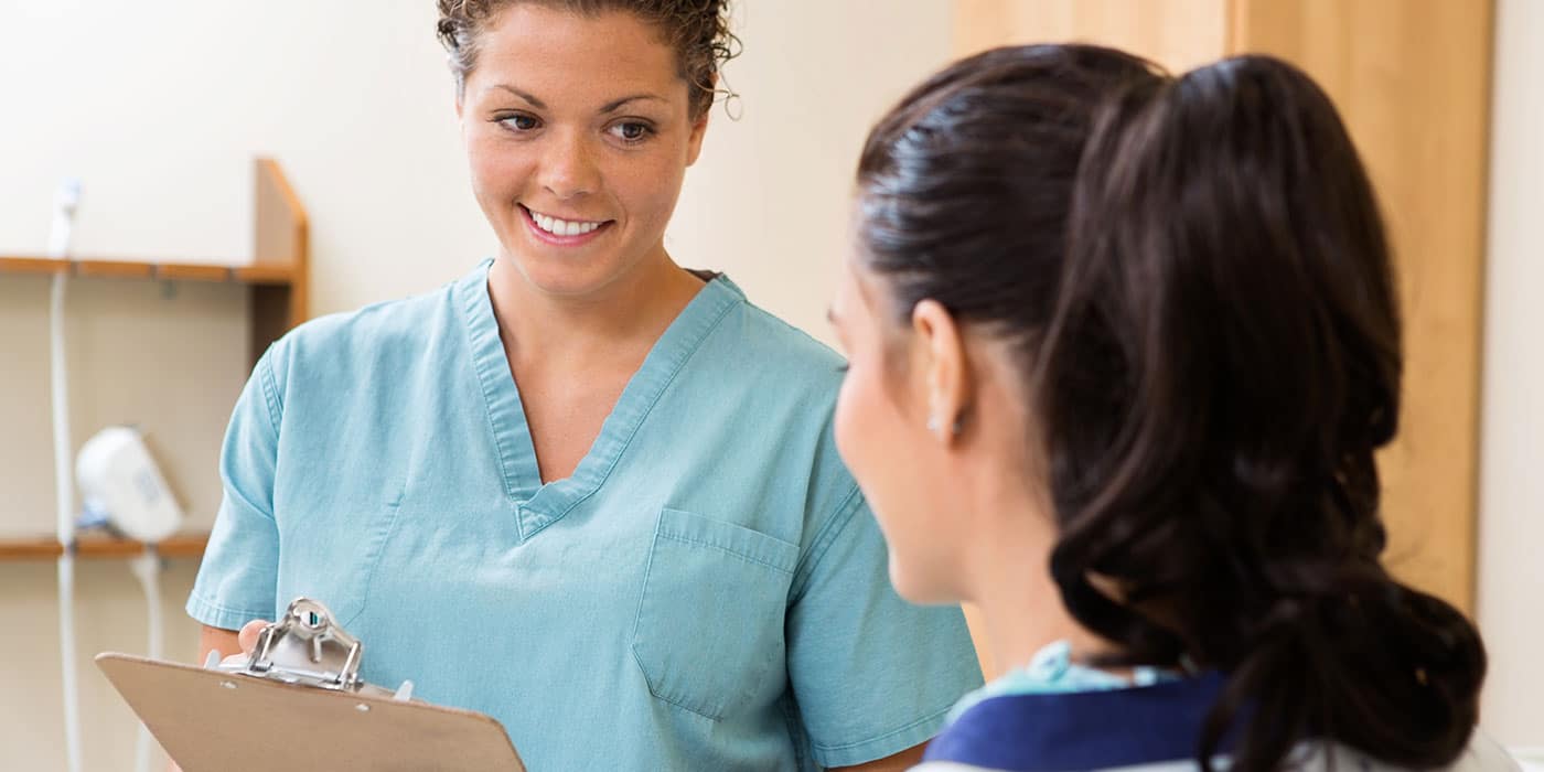 An instructor in scrubs stands with a clipboard smiling at an onlooking student.