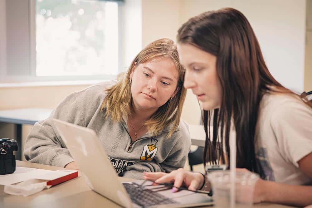 Learn by Doing or Experiential Learning, is demonstrated as one student leans in to see how another is using a program on a laptop.