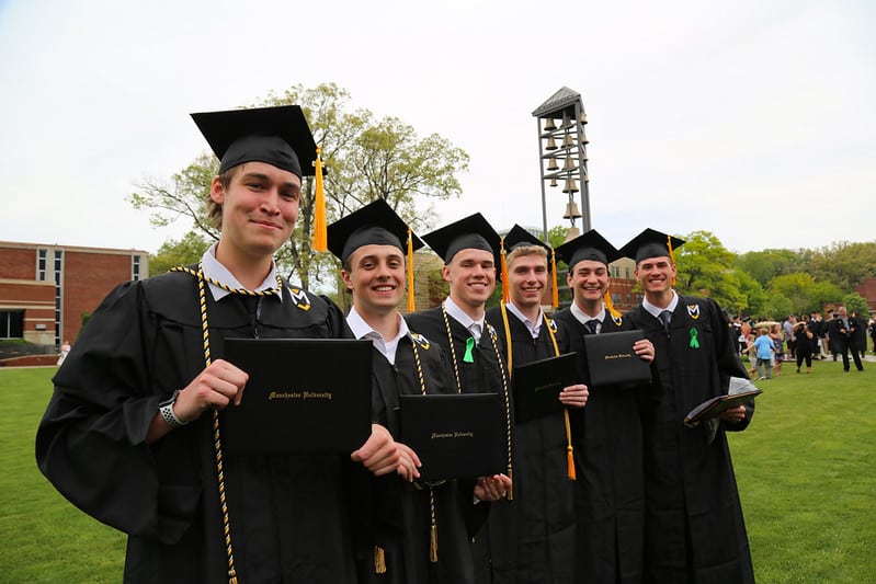 6 men graduates stand together in front of the chime tower after graduation