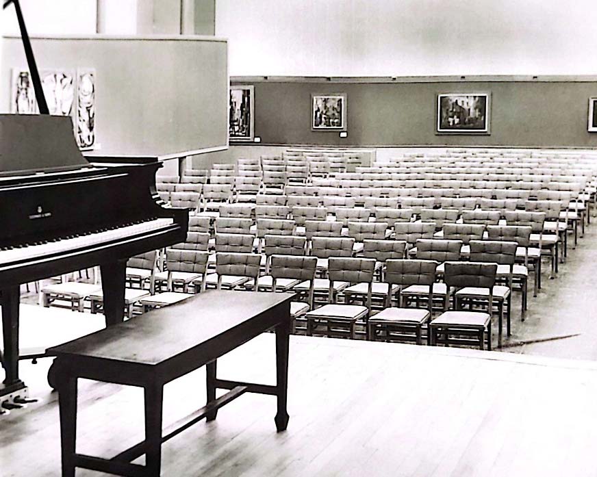 Original auditorium space used in the original administration building with a bell tower. Displays a piano on stage in front of empty seats