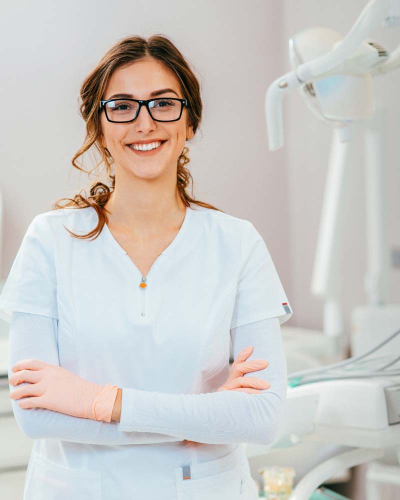 A female dentist stands in her office with chair and lamp behind her.