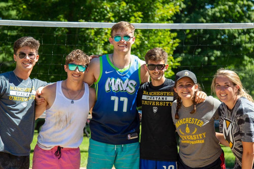 6 friends pose on the sand volleyball court on a sunny day.