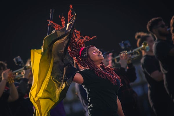 A female student of color wearing pink makeup twirls a golden flag