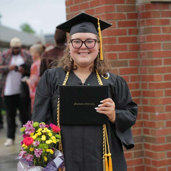 A grad stands in front of the chime tower holding a bouquet of flowers.