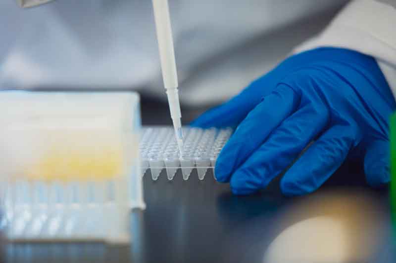 LabCloseUp Image displays a close up shot of a blue gloved hand and lab materials on a black surface.
