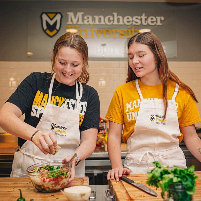 2 female students wearing MU aprons mix a salsa in the NGx lab.