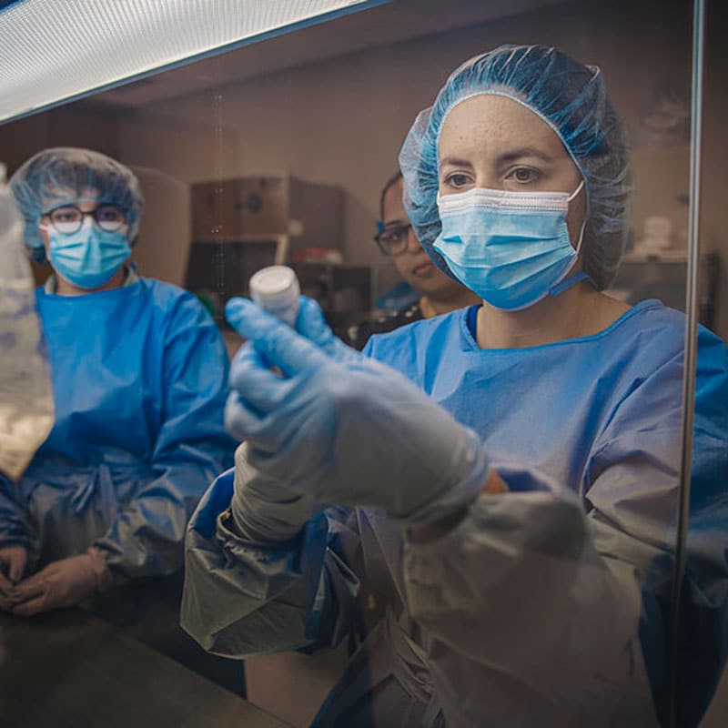 PharmdNeedle wearing full blue scrubs, mask, head covering, and gloves, a student uses real-world techniques in the lab.
