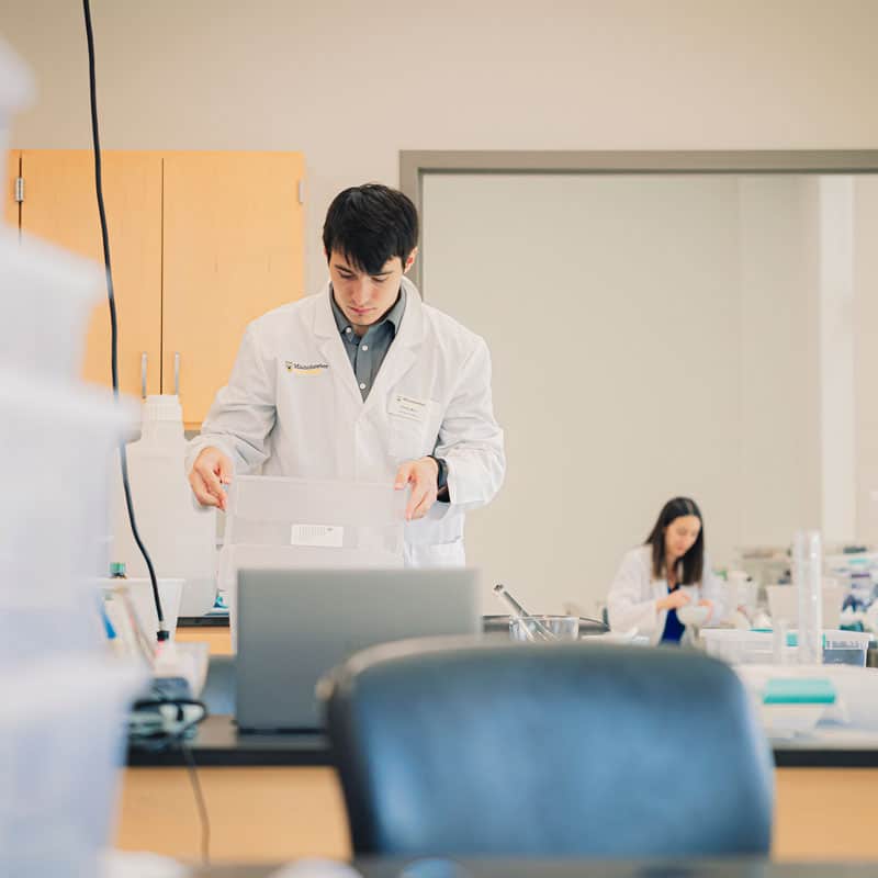 StackBinsPharm Wearing a white lab coat, a student in the pharmacy lab stacks bins.